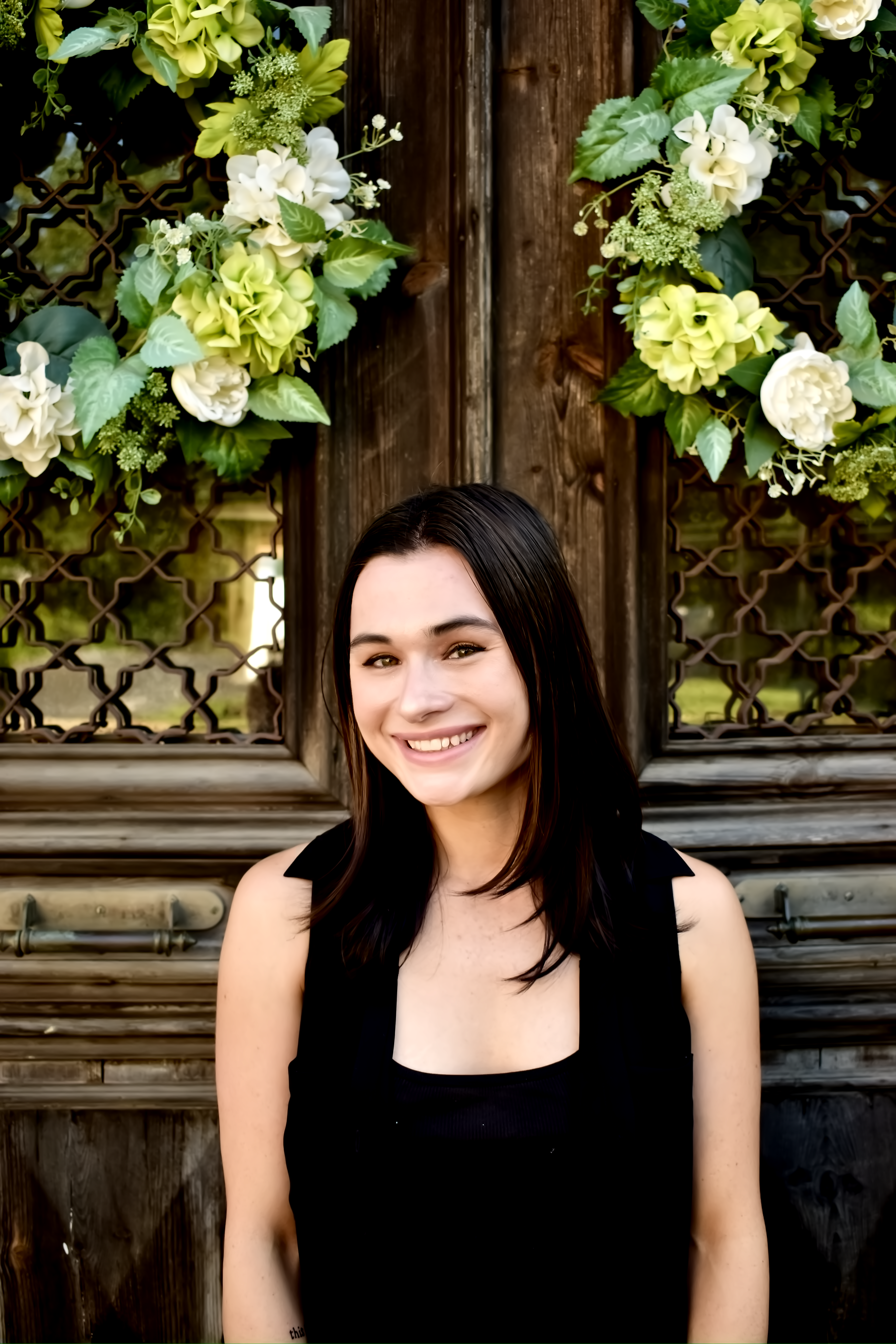 Darby Pistilli is standing in front of a brown door with flowers with a black shirt on, she is smiling at the camera